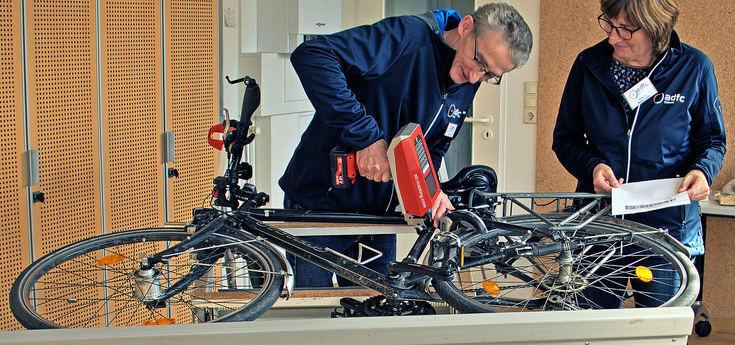 Ein Mann codiert mit einem Nadeldrucker ein auf der Seite liegendes Fahrrad, während die Besitzerin zuschaut. 